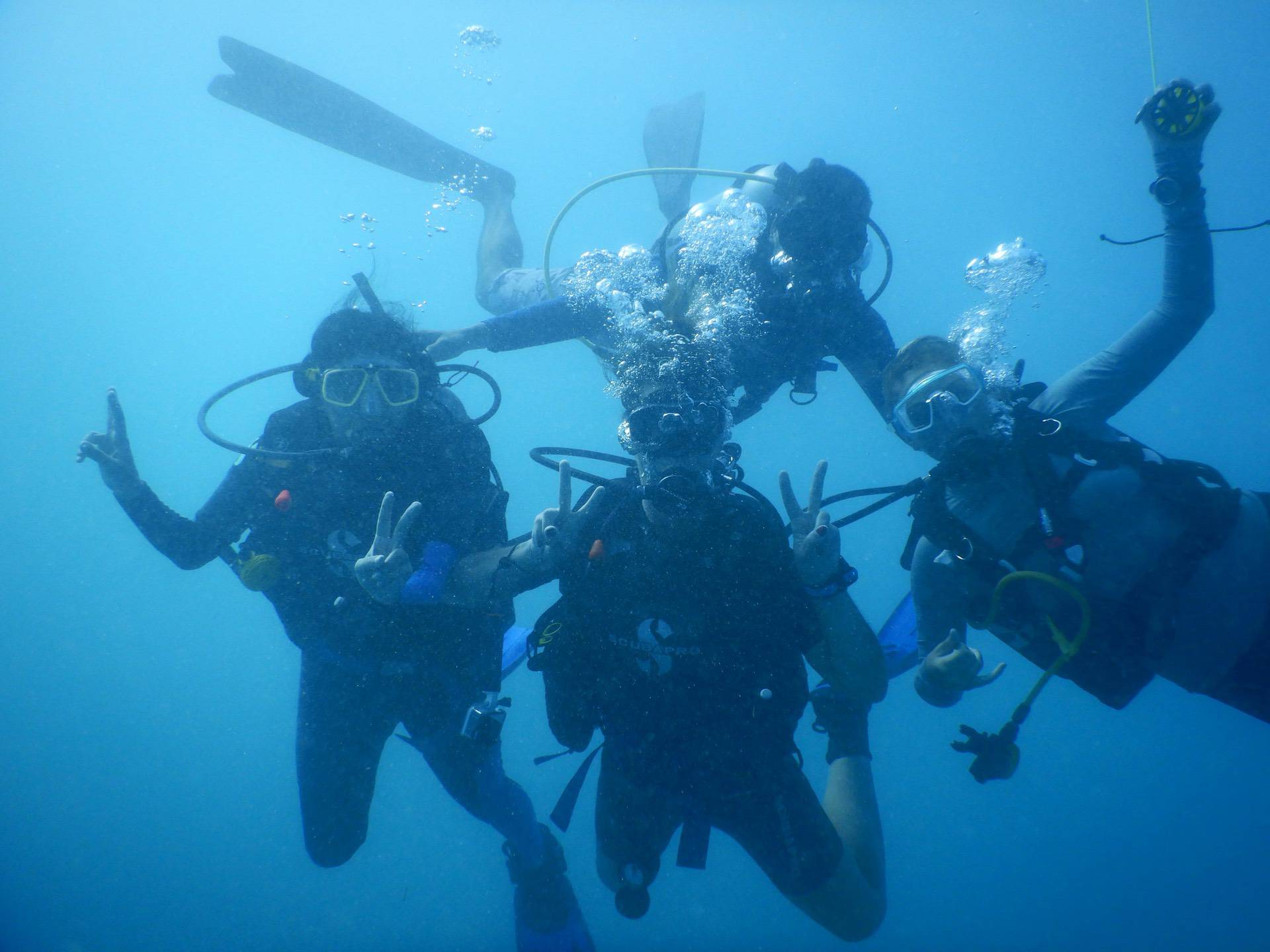 Four scuba divers underwater, wearing masks and breathing apparatus, pose while making peace signs with their hands. Bubbles rise from their equipment.