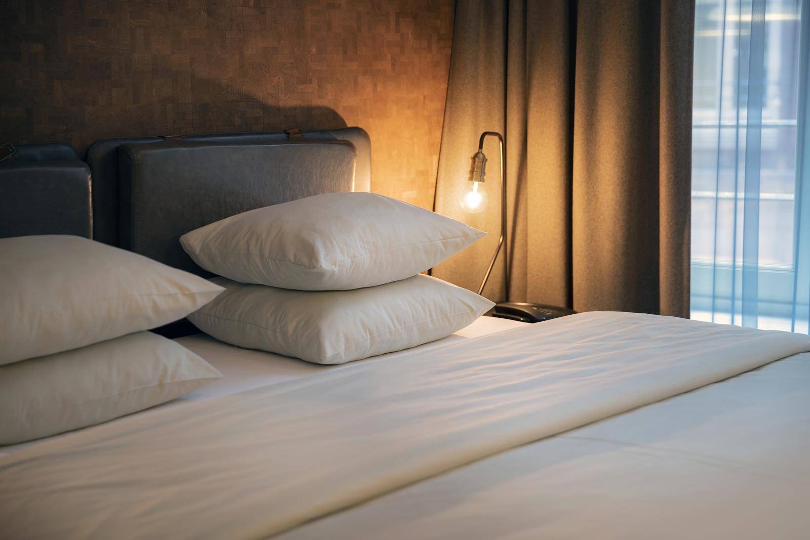 A neatly made bed with two stacked pillows on each side. A single light bulb lamp is on a small table near the bed, with curtains drawn in the background.