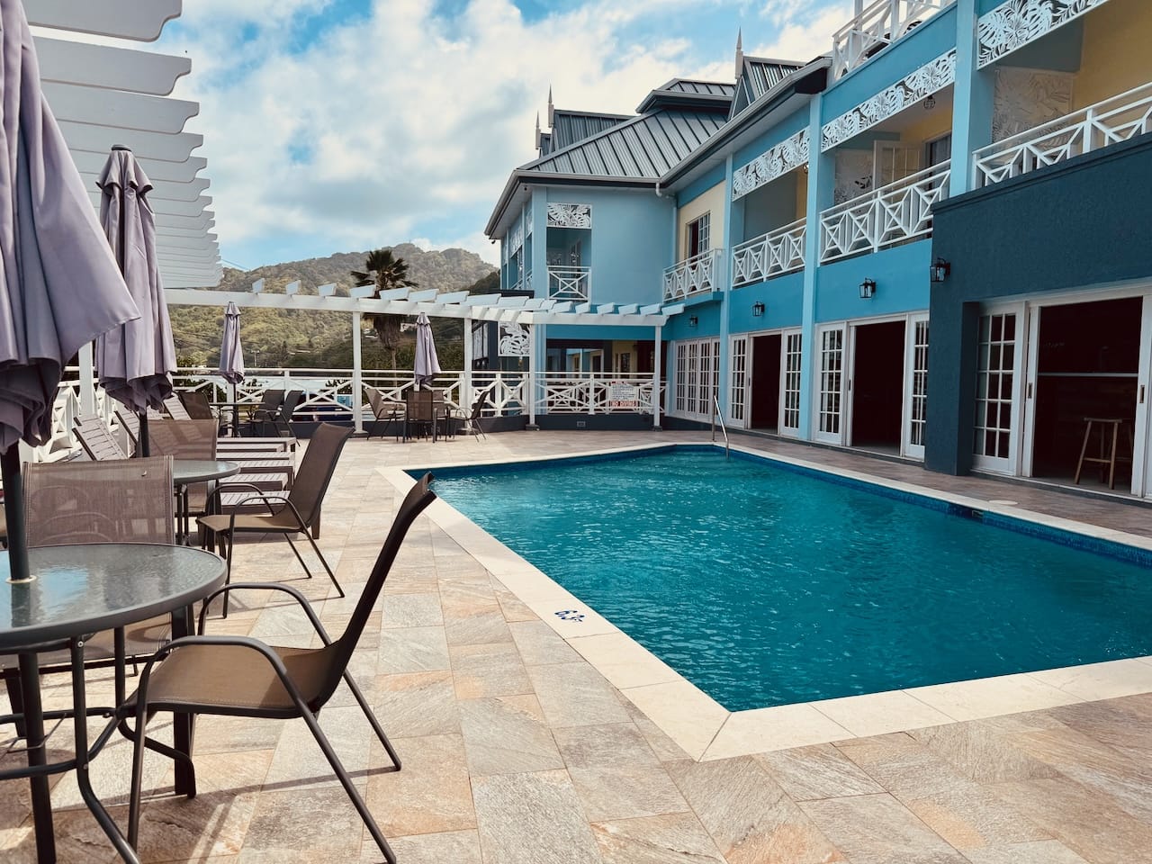 Outdoor pool area of a blue two-story building with lounge chairs, tables, and umbrellas on a sunlit day. Mountains are visible in the background.